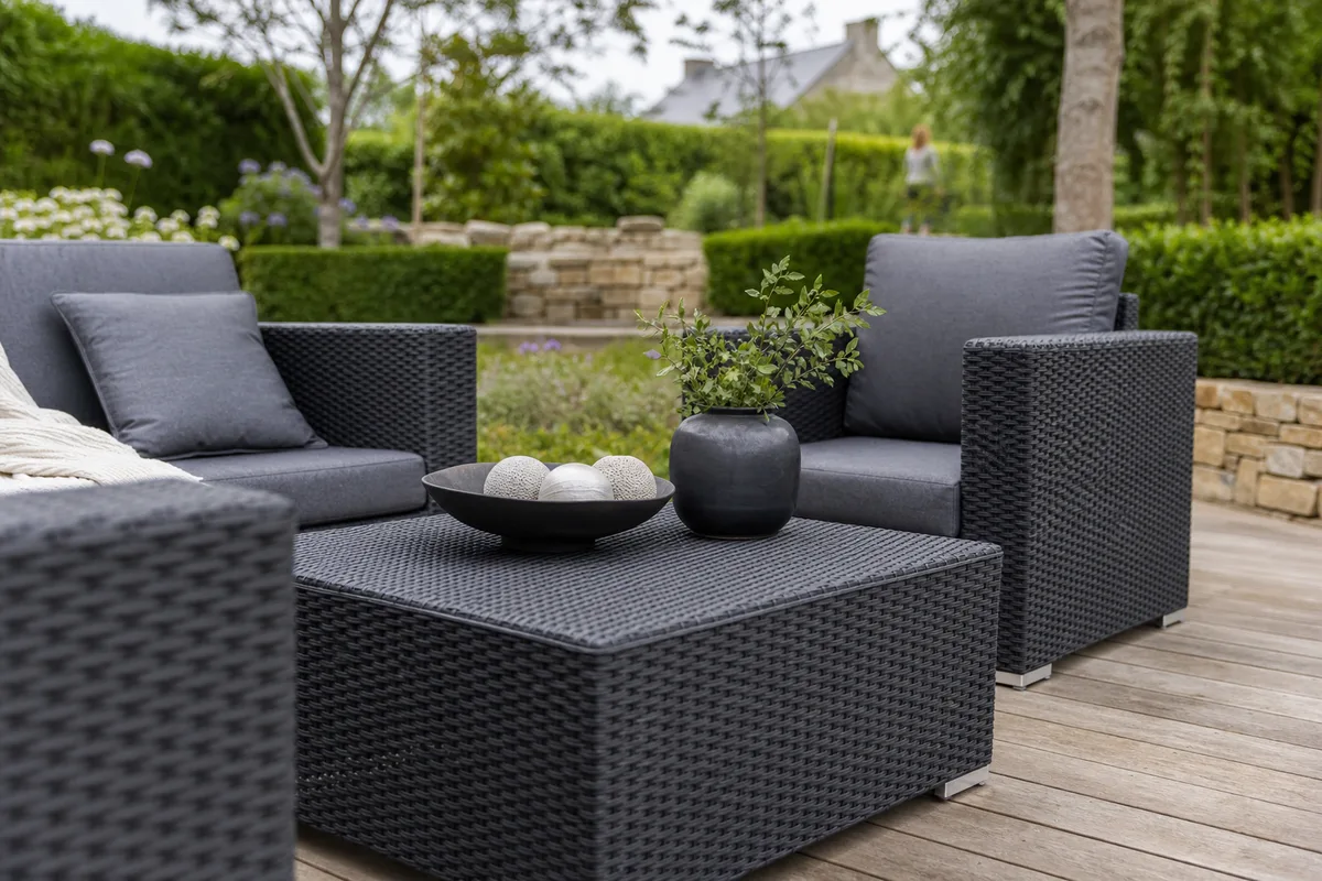 Table basse et fauteuils en résine tressée gris anthracite sur une terrasse en bois exotique, jardin structuré en arrière-plan avec haies et pierre naturelle, lumière douce de fin d’après-midi côté Lorient