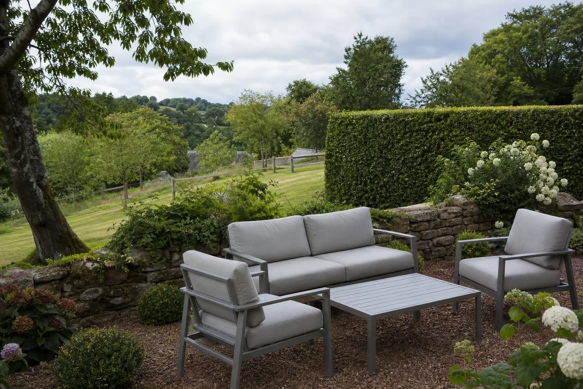 Coin salon extérieur avec fauteuils en aluminium gris et coussins déhoussables, abrité par une haie taillée et un muret en pierre, jardin en pente douce typique du pays de Quimperlé